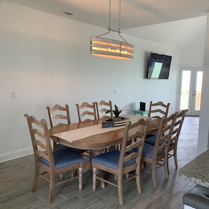 Uma área de refeições com mesa e cadeiras de madeira, um lustre pendurado, uma TV de tela plana na parede e uma porta que dá para uma área externa.