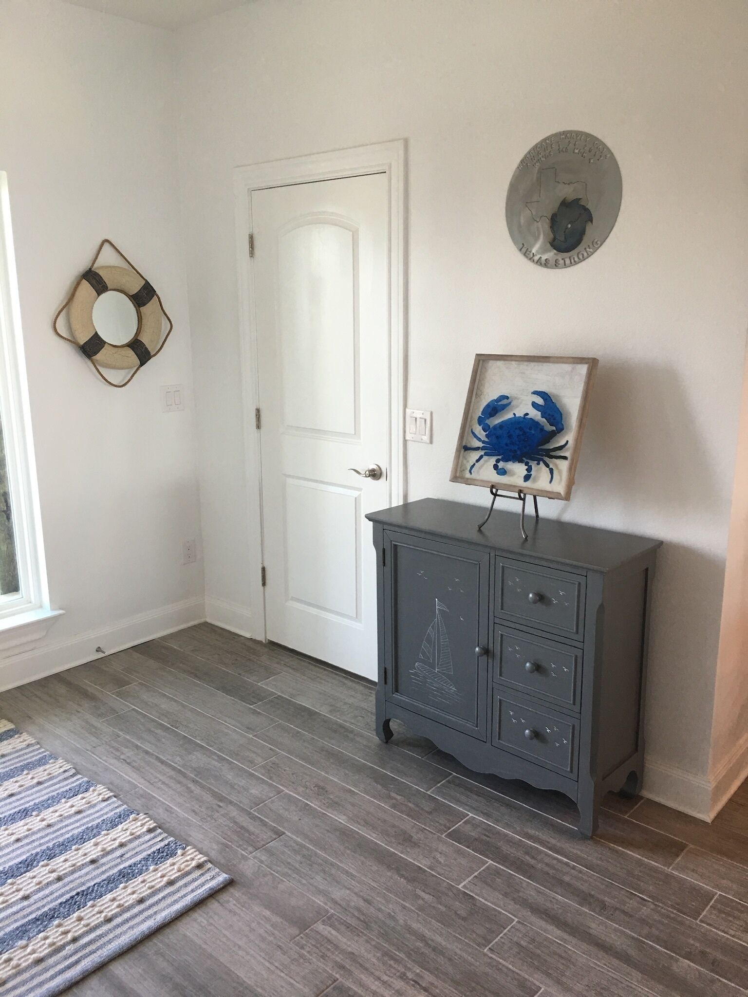 A room with a white door, a gray cabinet with drawers, a blue crab painting, a life ring, and a Texas state plate on the wall.