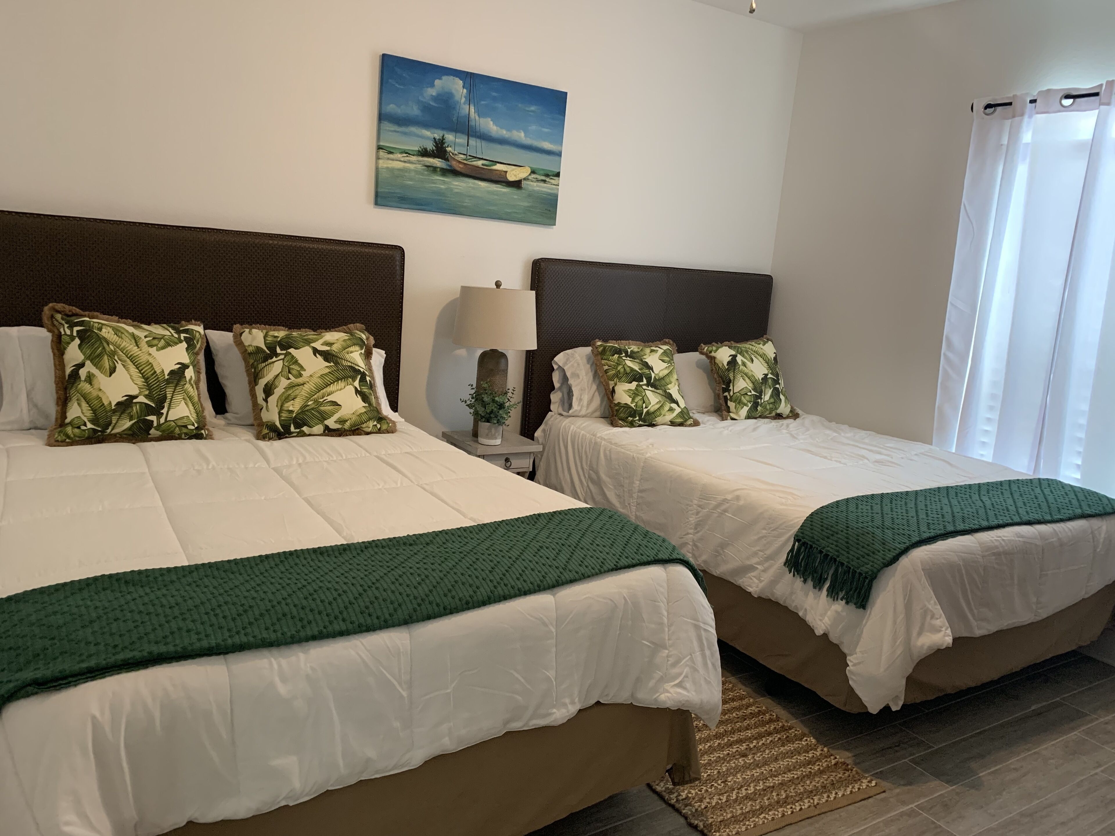 Camera d'hotel con due letti, ognuno con biancheria da letto bianca e coperte verdi. Si nota un quadro raffigurante una barca sulla parete, una lampada sul comodino e una finestra con tende bianche.