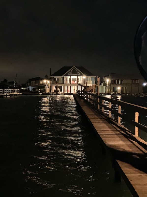 Uma propriedade à beira-mar com um cais de madeira que se estende por águas calmas, uma casa grande com várias janelas e uma varanda, e um edifício menor ao lado da casa principal.