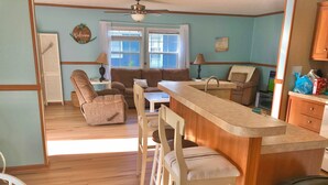 A cozy room with a wooden floor, a sofa, a recliner, a dining area with a bar, and a window with curtains. - Beautiful Beach Getaway! Walk to Beach! Sleeps 6. (Oak Island)