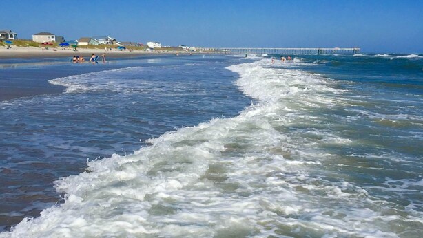 A beach with waves, a pier, and houses in the background. - Beautiful Beach Getaway! Walk to Beach! Sleeps 6. (Oak Island)