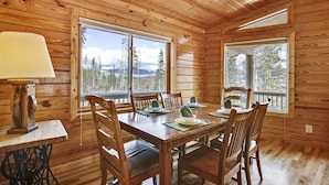 Eine Holzhütte mit Essbereich, inklusive Tisch für vier Personen, Holzsesseln und großen Fenstern mit Blick auf Bäume und Berge.