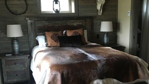 Un dormitorio rústico de madera con una cama grande, dos lámparas de noche, un ventilador de techo, una decoración con cráneo de vaca y una ventana que proporciona luz natural.