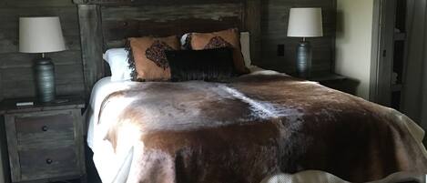 Un dormitorio rústico de madera con una cama grande, dos lámparas de noche, un ventilador de techo, una decoración con cráneo de vaca y una ventana que proporciona luz natural.