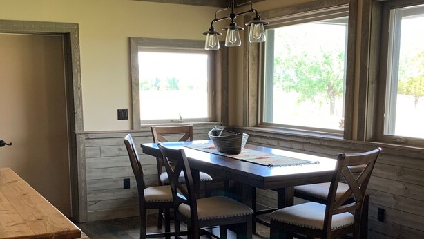 Un comedor con mesa y sillas de madera, lámpara colgante y ventanas amplias con vista a follaje.