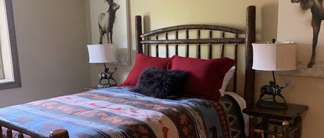 Una cama con cabecera de madera, colcha estampada y almohadas rojas. Se observa un mesita de noche con una lámpara y un objeto decorativo.