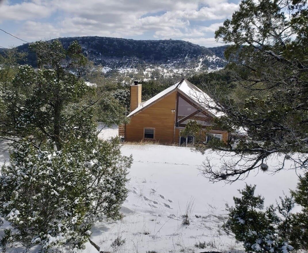 A house with a sloped roof, surrounded by snow-covered ground and trees, with a mountainous backdrop.