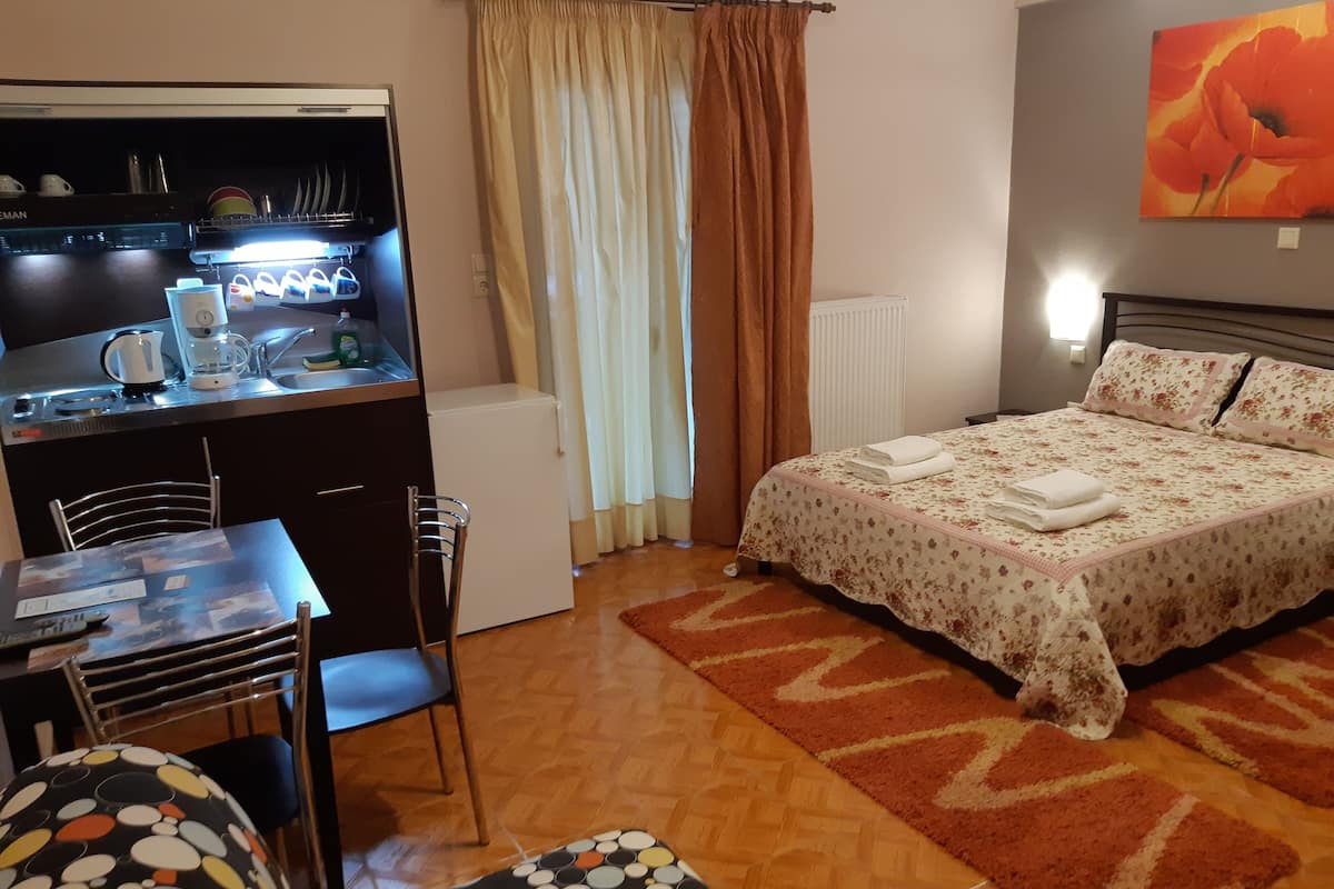 A compact hotel room with a kitchenette, a bed with a floral bedspread, a dining area with a chair, and a colorful rug.
