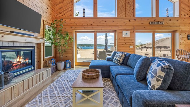 A cozy cabin interior with a fireplace, large windows, a blue sofa, and a view of the beach. - Lake frontage, Dune beach sand from deck to water! One of a kind. (Mears)
