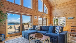 A wooden cabin interior with a large sofa, a coffee table, and a view of a beach. - Lake frontage, Dune beach sand from deck to water! One of a kind. (Mears)