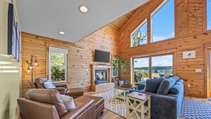 A cozy cabin interior with a fireplace, leather recliner, sofa, and large windows offering a view. - Lake frontage, Dune beach sand from deck to water! One of a kind. (Mears)