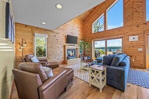 Interior acogedor de una cabaña con chimenea, sillón de cuero, sofá y ventanas amplias con vistas.