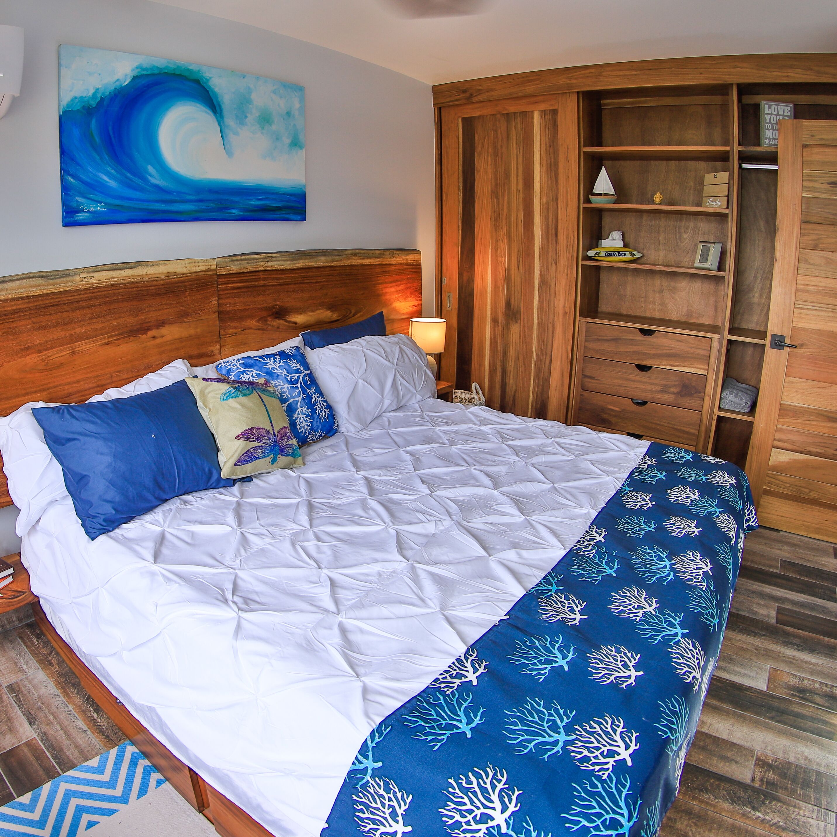 Un dormitorio con cabecera de madera, cama con ropa de cama blanca y azul, un armario de madera y un cuadro de una ola en la pared.