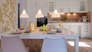 A modern kitchen with white cabinetry, pendant lighting, and a breakfast setup with plates, cups, and a fruit bowl. - Tide App. 3 gold right (Ostseebad Boltenhagen)
