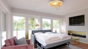 A modern bedroom with a bed, a pink armchair, a round coffee table, and a TV mounted on the wall. - Tide App. 3 gold right (Ostseebad Boltenhagen)