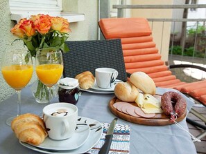 A breakfast setup with croissants, pastries, ham, cheese, and a sausage on a wooden board, along with two glasses of orange juice and a cup of coffee on a table.