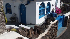 Un edificio blanco con puertas y ventanas azules, techo de tejas y un muro de piedra en primer plano.