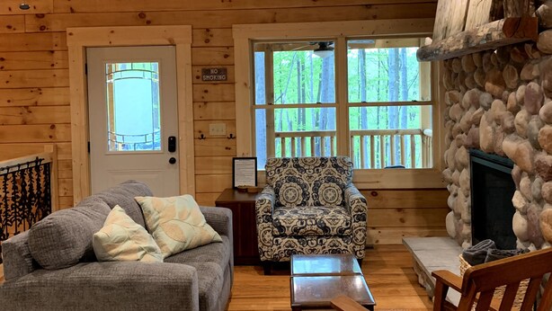 A cozy cabin interior with a wooden ceiling, a stone fireplace, a sofa, a patterned armchair, and a dining table. - Incredible secluded cabin featuring amazing forest views off of the back deck! (Laurelville)
