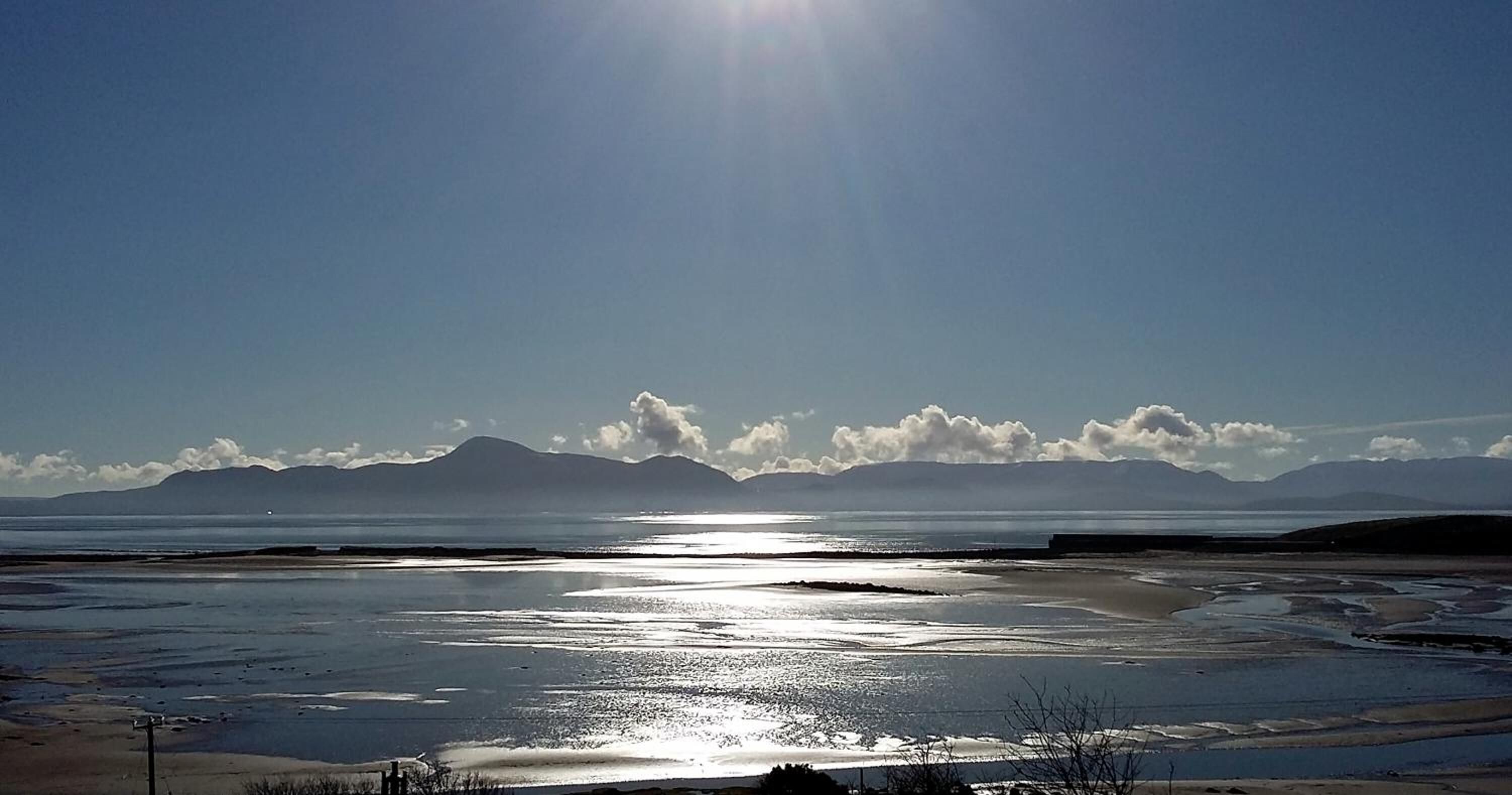 A beach with a clear sky, mountains in the distance, and a calm sea reflecting the sunlight.