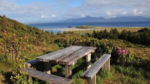 Een houten picknicktafel met bankjes in een grasveld, met uitzicht op de oceaan en bergen in de verte.