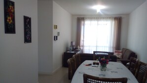 A dining area with a table set for four, surrounded by wicker chairs. There is a vase with flowers on the table, and a television is visible in the background. - BEAUTIFUL AND COMFORTABLE 3QTS APARTMENT ON THE BEACH OF ENGLISH IN 180 MTS FROM THE BEACH (Ingleses do Rio Vermelho)