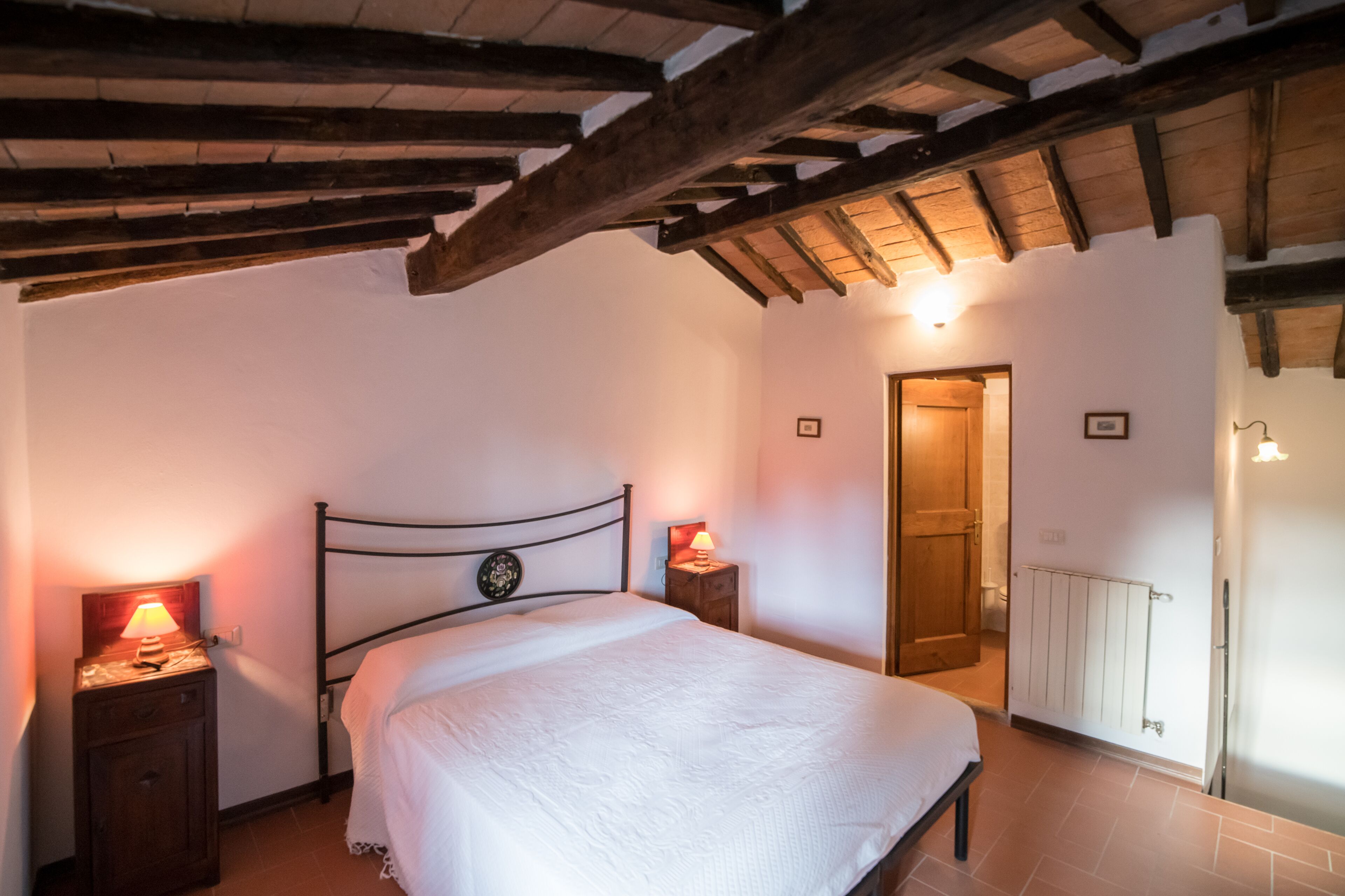 A bedroom with a wooden ceiling, a bed with white bedding, two bedside tables with lamps, and a radiator on the wall.