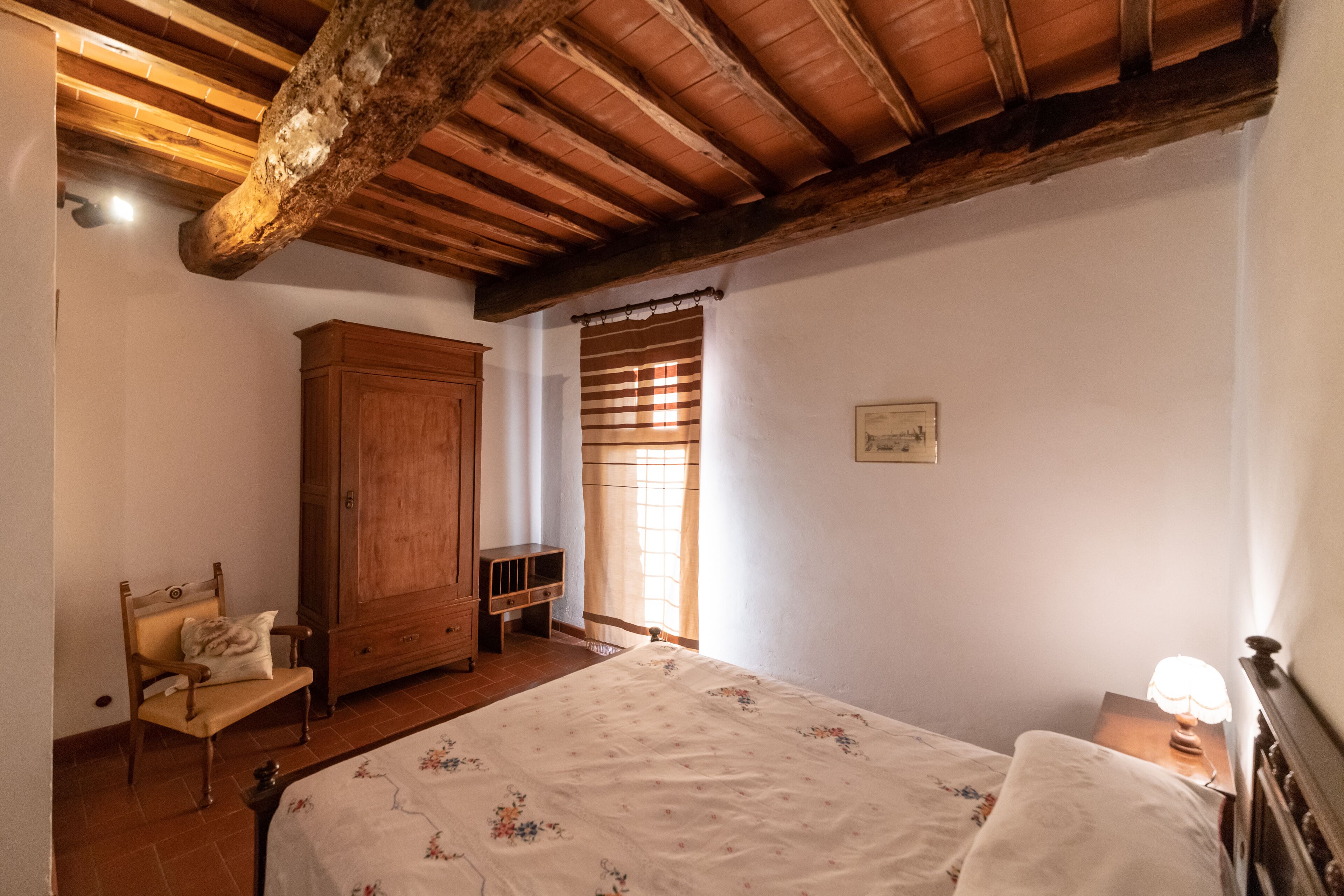 A bedroom with a wooden ceiling, a bed with a floral bedspread, a wooden wardrobe, a chair, and a window with curtains.