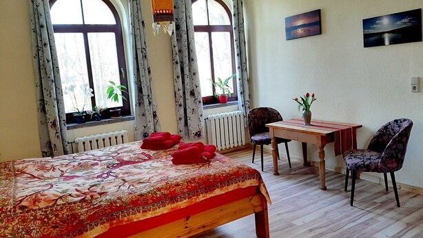 A room with a bed, a table, and two chairs. There are windows with curtains and potted plants on the windowsill. - Vacation apartment at the Stadtpark_Seerose - Holiday apartments at the Stadtpark (Lutherstadt Wittenberg)