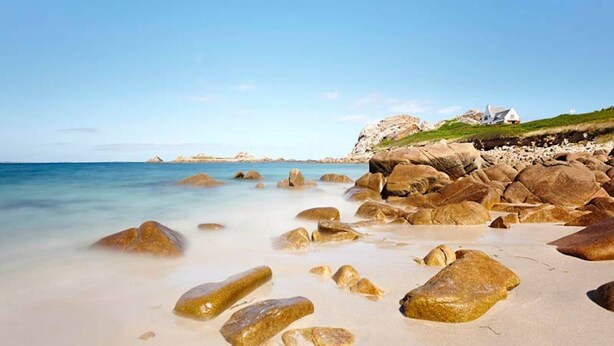 A beach with large rocks and a white building on a grassy hill.