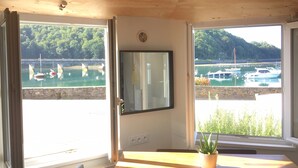A room with a wooden ceiling, a large window with a view of boats and greenery, a wall-mounted TV, and a potted plant on a wooden table.