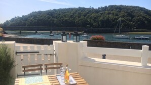 A terrace with a table set for two, overlooking a marina with boats and a forested hill in the background.