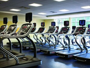 Een rij loopbanden met digitale displays, geplaatst in een sportschool met een blauwe vloer en ramen op de achtergrond.