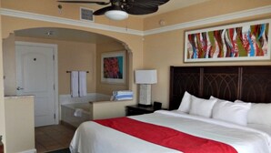 Habitación de hotel con una cama grande, ventilador de techo, baño con toallero y un cuadro en la pared.
