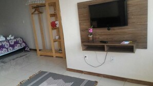 A living room with a flat-screen TV mounted on a wooden wall, a wooden shelf below it, and a tiled floor.