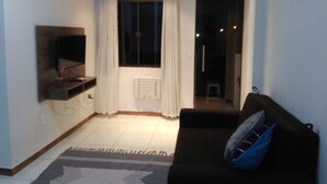 A living room with a dark sofa, a flat-screen TV mounted on the wall, a window with white curtains, and a small table with a vase.