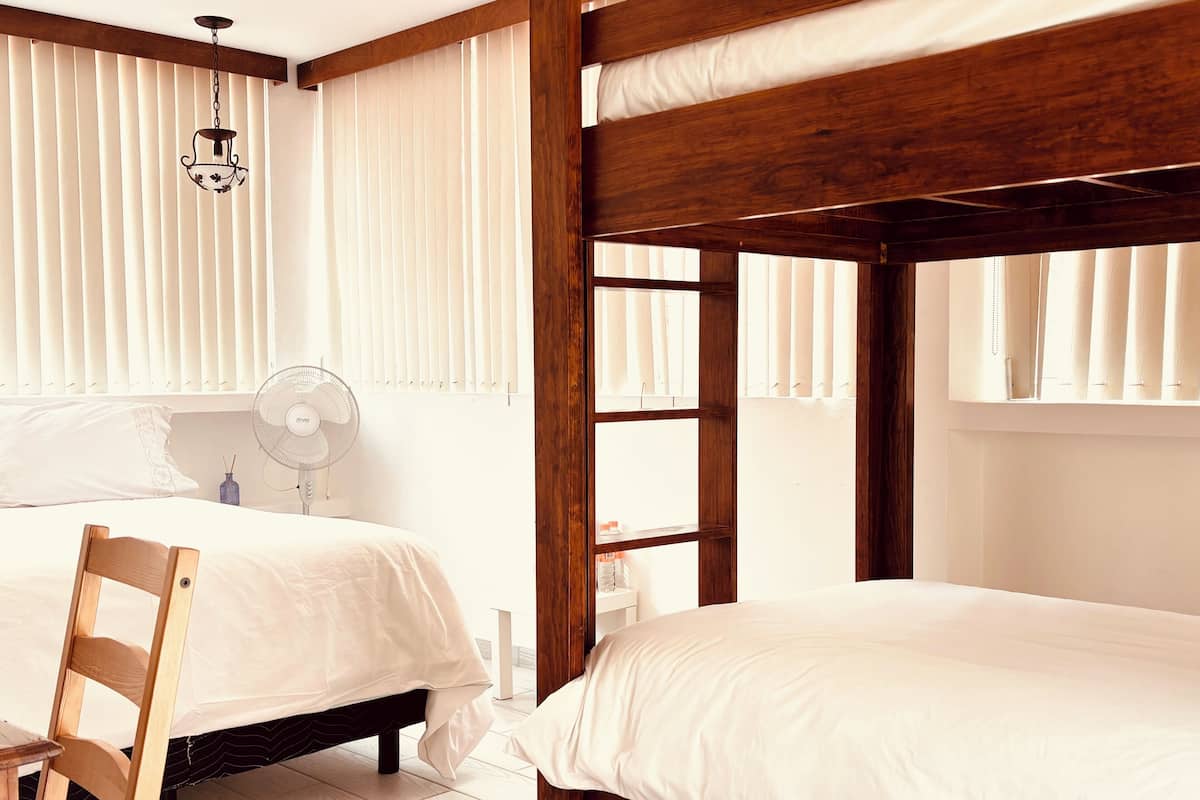 Een slaapkamer met twee houten stapelbedden, een nachtkastje, een stoel, een hanglamp en een raam met jaloezieën.