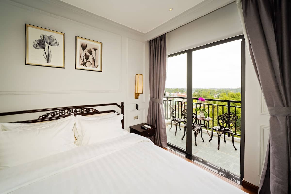 A hotel room with a large bed, two framed floral artworks on the wall, a nightstand, and a balcony with outdoor furniture.