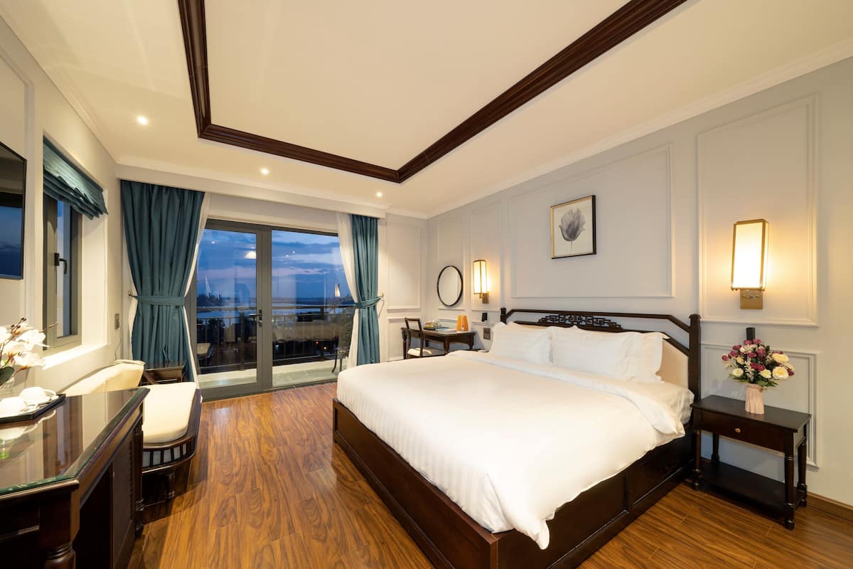 A hotel room with a large bed, a desk with a chair, a desk lamp, a nightstand with a vase of flowers, and a view of the city through the window.