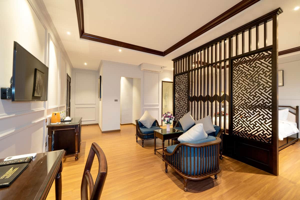 A hotel room with a wooden floor, a dining table with chairs, a sofa with cushions, a TV mounted on the wall, and a decorative partition with a geometric pattern.