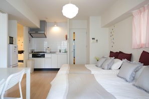 A modern, well-lit kitchen with white cabinets, a built-in oven, and a microwave. There is a dining area with a white table and chairs. The room features a large bed with white bedding and multiple pillows.
