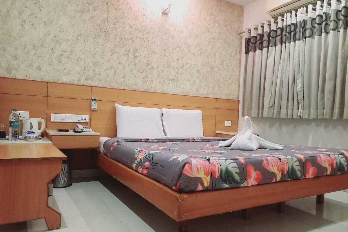 A hotel room with a wooden bed, floral bedspread, a nightstand with a kettle, and a window with curtains.