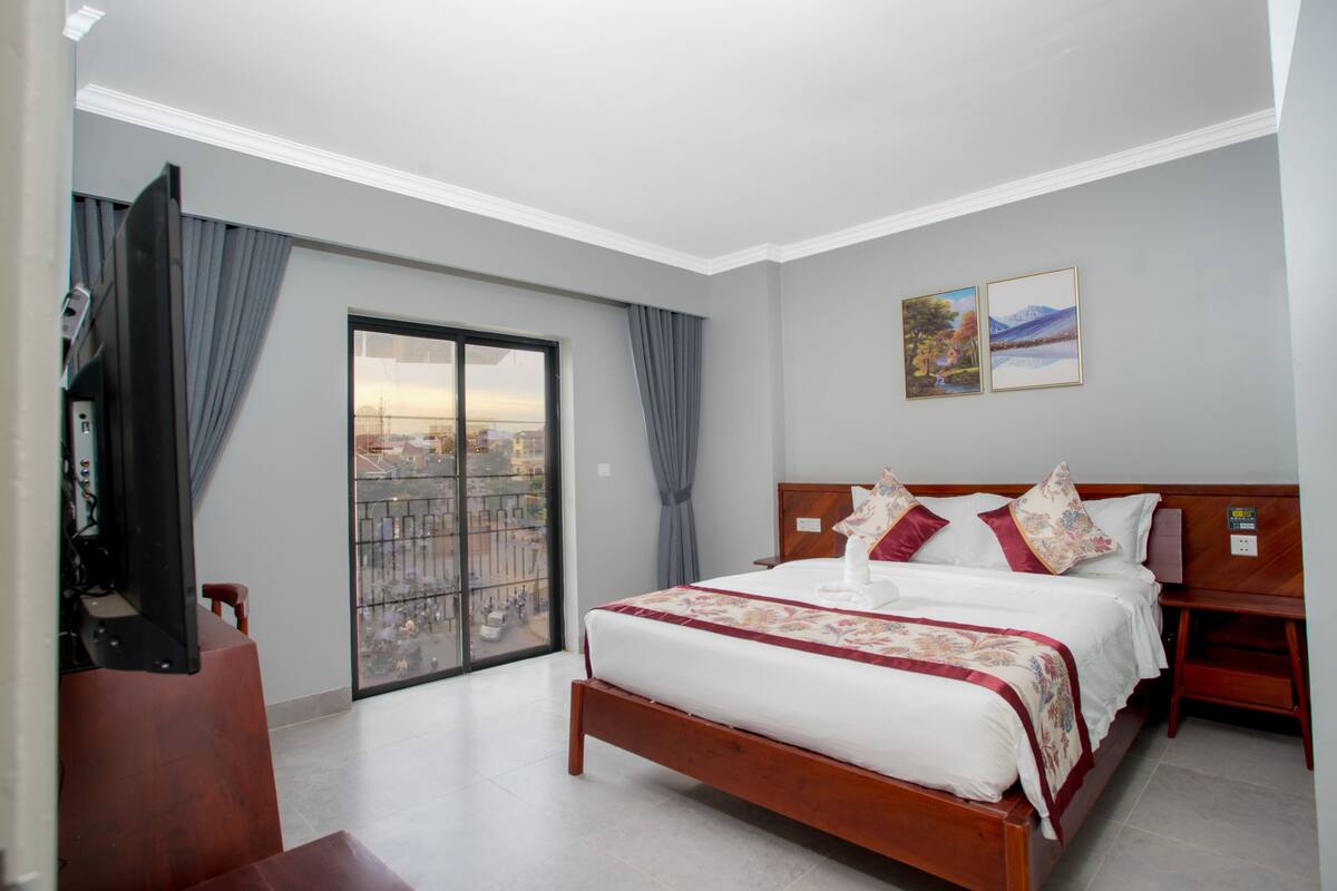 Habitació d'hotel amb un llit gran, una tauleta de nit de fusta, televisió i un balcó amb vista a la ciutat.