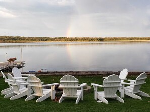 A lakeside view with four Adirondack chairs facing a calm lake, a dock with a dog, and a rainbow in the distance.
