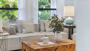 Ein Wohnzimmer mit einem Holztisch, einer weißen Vase mit Grünpflanzen, einem beigefarbenen Sofa und einer Lampe auf einem Beistelltisch.