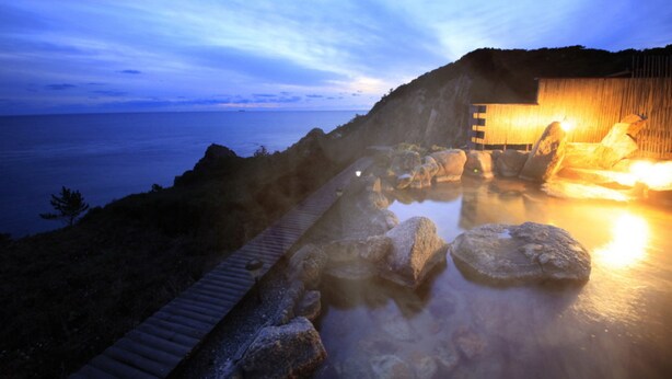 A hot spring area with a wooden deck, large rocks, and a building with warm lighting. - TheMana Village (Tosashimizu)