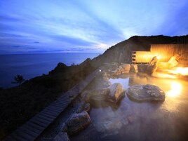 Un área de aguas termales con una terraza de madera, rocas grandes y un edificio con iluminación cálida.