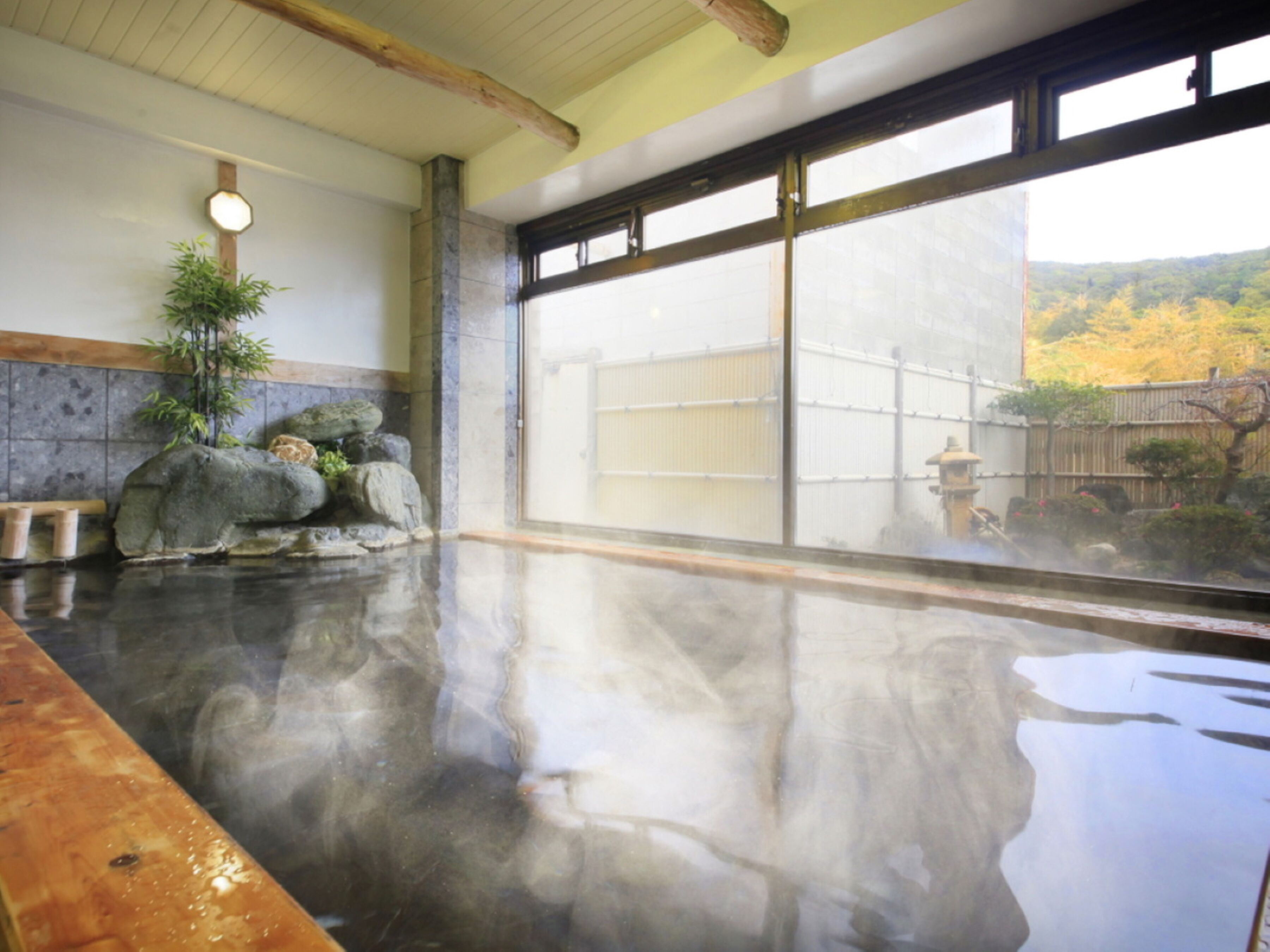 Un baño de aguas termales con un banco de madera, un arreglo de rocas de piedra y vistas a un jardín a través de grandes ventanales.