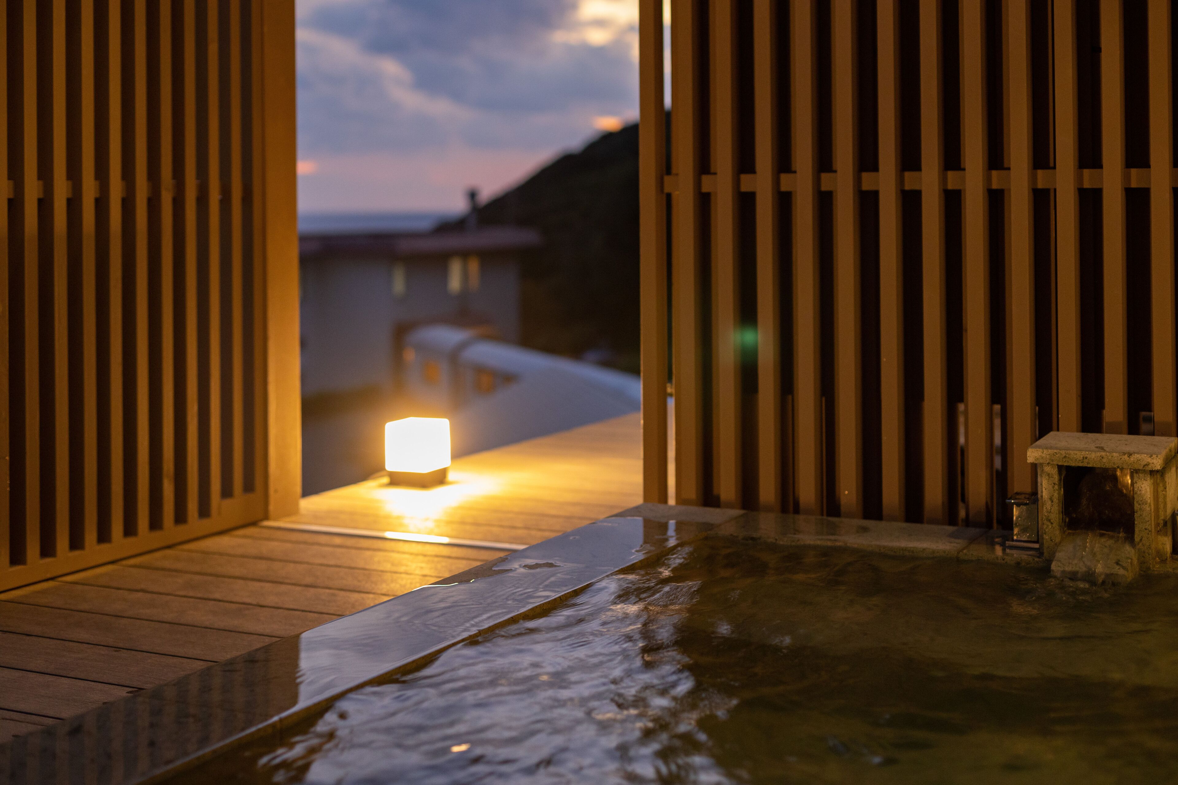 Una terraza de madera que conduce a un área de baño de piedra con una vela encendida, rodeada de listones verticales de madera.
