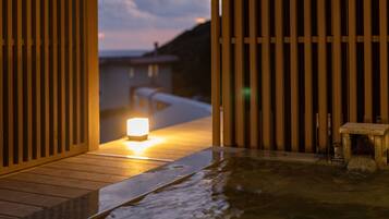 Una terraza de madera que conduce a un área de baño de piedra con una vela encendida, rodeada de listones verticales de madera.